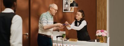 Senior guy at front desk doing check in routine in expensive five star hotel lobby. Traveller signing registration forms at front desk, checking room service and ammenities on holiday trip.